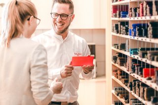 Ein lachender Optiker, der ein rotes Farbmuster in der Hand hält, berät eine Kundin. Im Hintergrund sind Regale mit weiteren Farbmustern zu sehen.