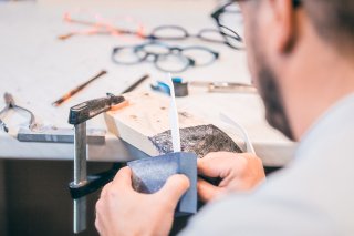 Ein Mann arbeitet in einer Werkstatt. Er hält ein weißes Brillengestell und feilt mit einem blauen Block daran, mit Fokus auf die Bearbeitung der Brille.