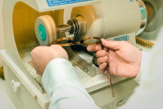 Detailaufnahme eines Optikers, der ein braunes Brillengestell mit beiden Händen an eine Poliermaschine hält, um es zu schleifen und zu polieren.