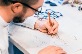 Ein bärtiger Mann, der eine Brille trägt, zeichnet einen Entwurf für eine Brille mit einem Bleistift auf Papier. Im Hintergrund liegen fertige Brillengestelle.