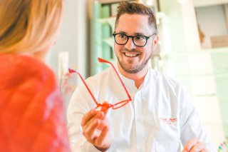 Ein freundlich lächelnder Optiker, der selbst eine Brille trägt, berät eine Kundin und präsentiert ihr ein rotes Brillengestell.