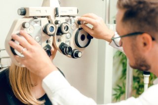 Der Brillenmacher führt mit einem Phoropter eine Sehtest durch, indem er die Rädchen des Geräts dreht. Eine blonde Patientin sitzt ihm gegenüber.