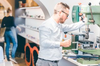 Ein Optiker arbeitet an einer grünen Werkzeugmaschine. Er trägt eine weiße Brille und ein weißes Hemd und konzentriert sich auf die Maschine.