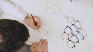 Von oben fotografiert. Eine Person zeichnet mit einem Bleistift Brillengestelle auf ein Blatt Papier. Auf dem Tisch liegen fertige Brillengestelle.